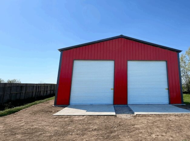 Red clear span building with two roll-up doors, set in a grassy area under a clear blue sky.