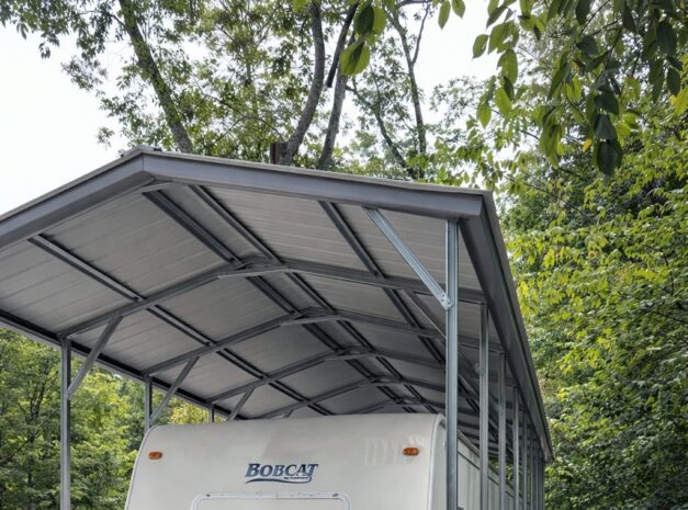 A-frame carport sheltering a travel trailer, surrounded by trees and gravel, showcasing durable construction.