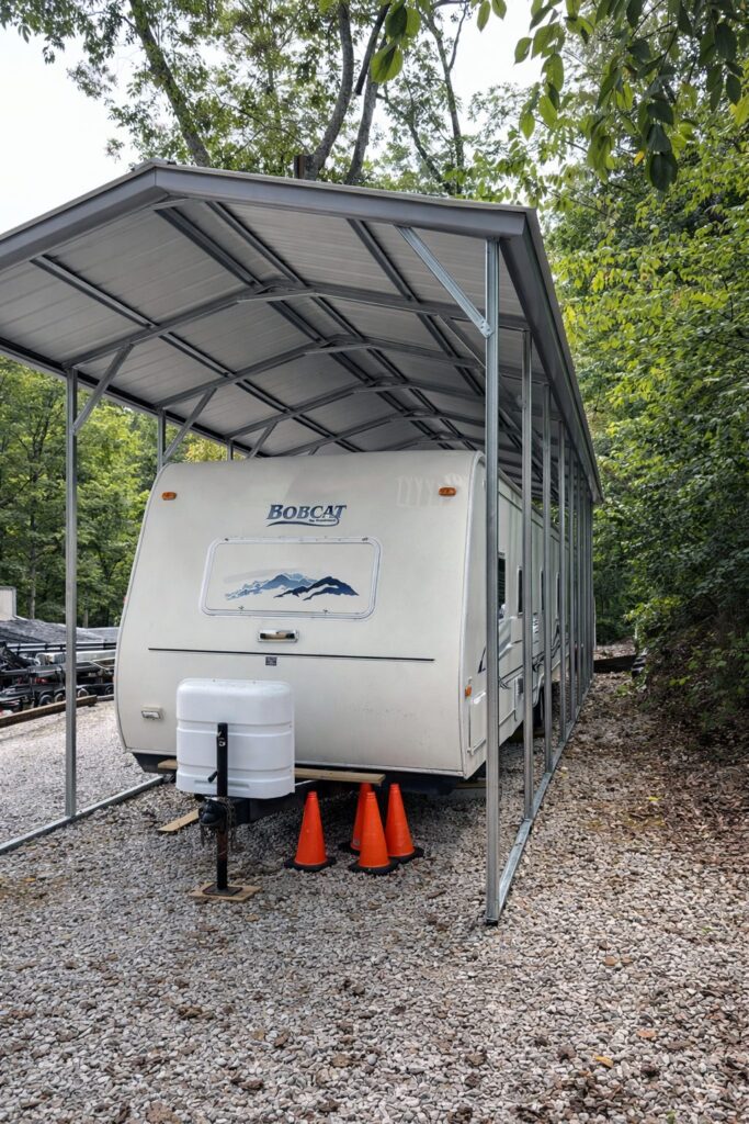 A-frame carport sheltering a travel trailer, surrounded by trees and gravel, showcasing durable construction.