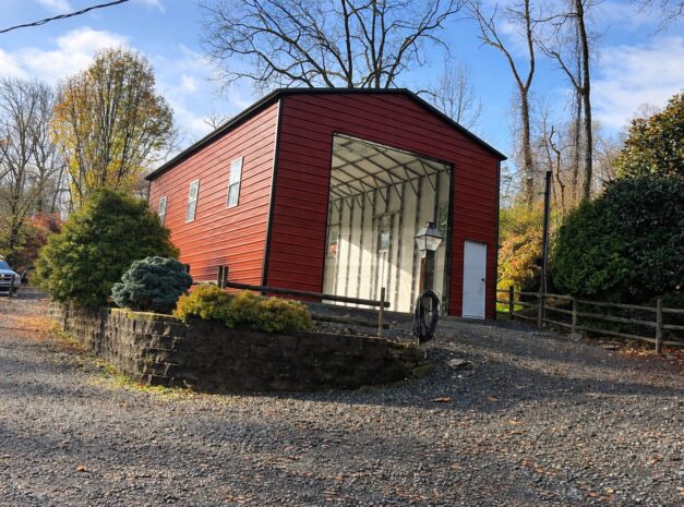 Custom RV carport with a red exterior, set in a scenic landscape with trees and a gravel driveway.