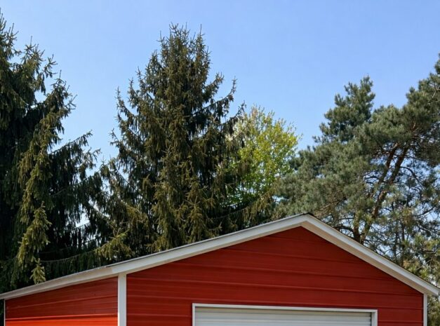 A red standard shed with a white roll-up door, set in a grassy area surrounded by trees.