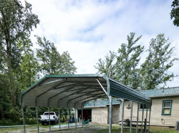 A standard carport structure with a metal frame, set on gravel and grass, near a house in Muskogee, OK.