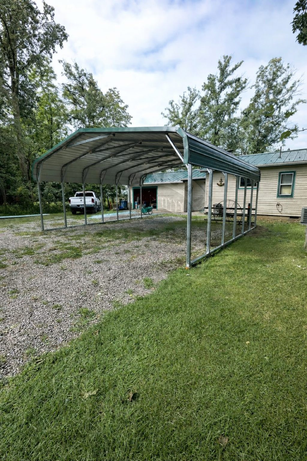 A standard carport structure with a metal frame, set on gravel and grass, near a house in Muskogee, OK.