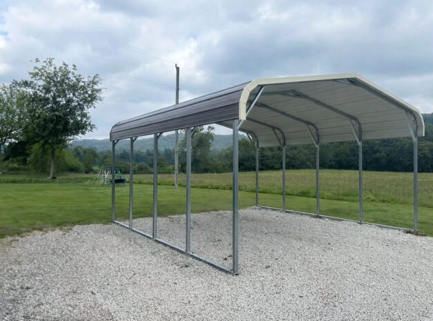 Standard carport with a metal frame and roof, set on gravel in a grassy area near trees.
