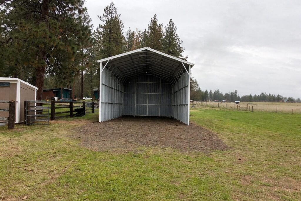 A spacious A-framed carport with a metal structure, set in a grassy area surrounded by trees.
