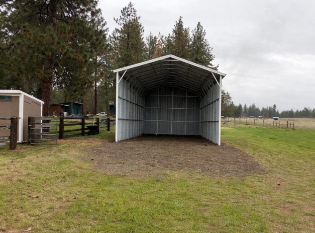A spacious A-framed carport with a metal structure, set in a grassy area surrounded by trees.