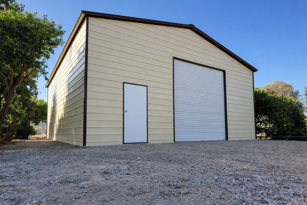 A side view of a tan A-framed carport with a roll-up door and a side entrance, surrounded by greenery.