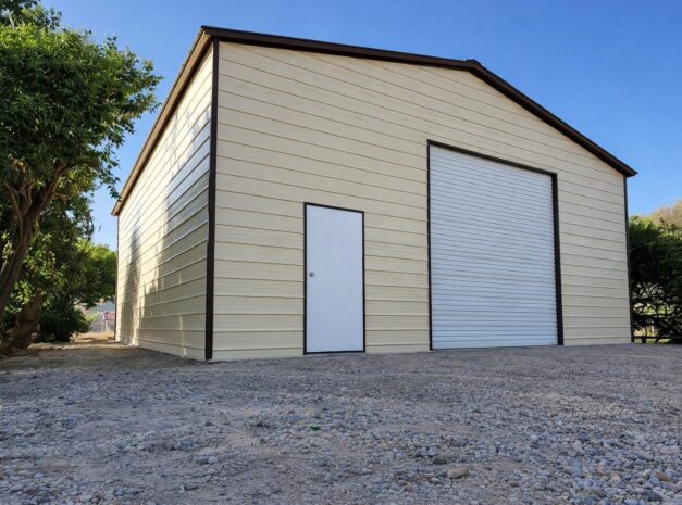 A side view of a tan A-framed carport with a roll-up door and a side entrance, surrounded by greenery.
